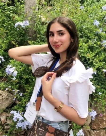 A young woman with long, dark brown hair smiling while standing in front of a garden of green plants and light purple flowers. She is wearing a white ruffled t-shirt, a gold watch, and a brown patterned crossbody bag with a small matching pouch.