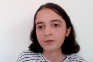 A young person with shoulder-length dark hair and a striped shirt looking directly at the camera. The headshot is taken against a plain, light-colored wall in a simple indoor setting.