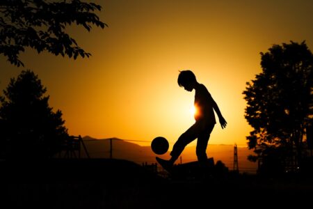 The silhouette of a young person kicking a soccer ball into the air against a vibrant orange and yellow sunset. Trees and distant mountains are visible in silhouette, and the sun is positioned directly behind the person, creating a glowing backlight effect.