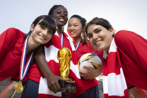 A low-angle close-up of four diverse, smiling female soccer players posing closely together after a victory. They wear red jerseys, gold winner's medals, and red-and-white striped scarves. Two players in the center hold a gold trophy resembling the World Cup, while another player holds a soccer ball.