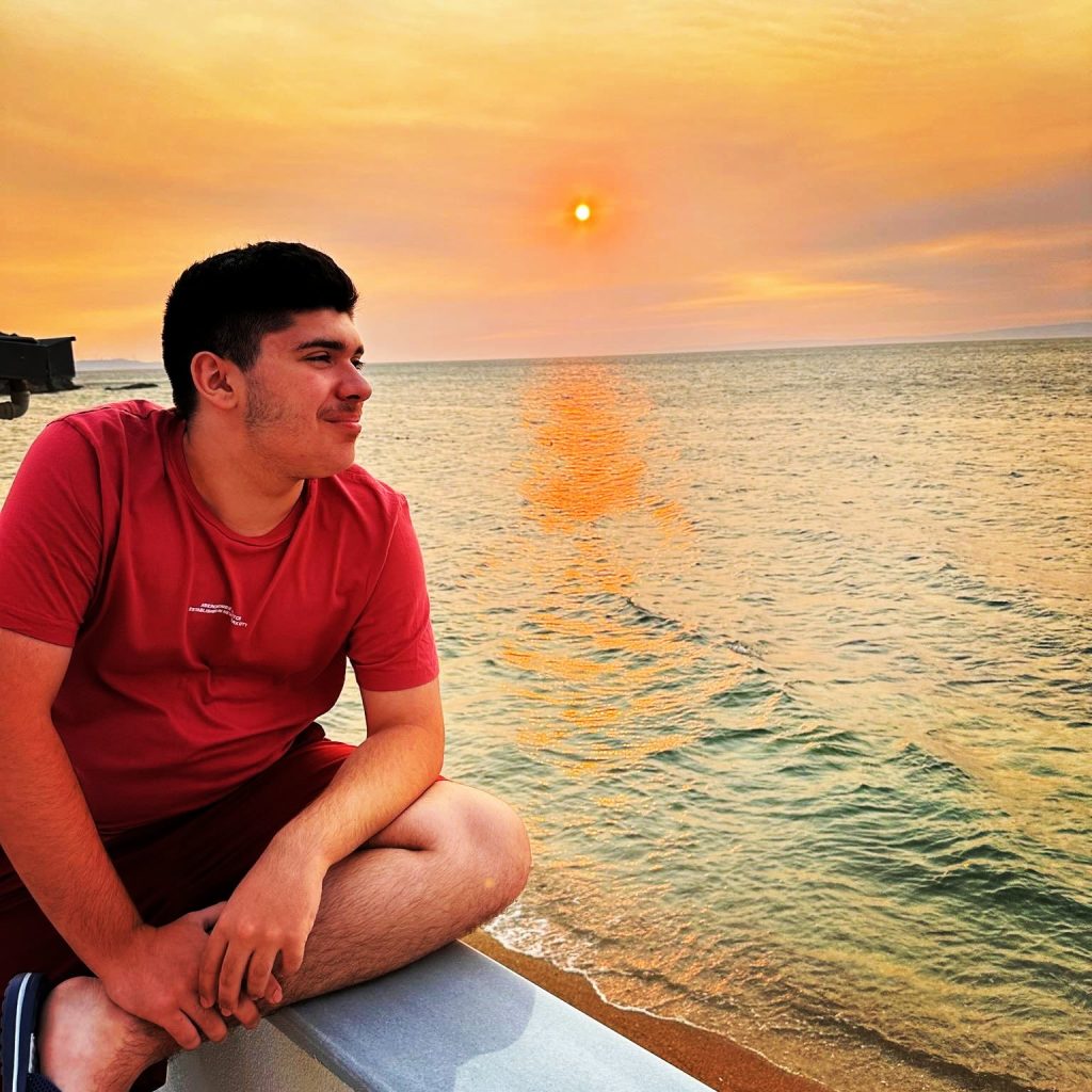 A young man in a red t-shirt sits on a ledge looking out over the ocean at sunset, with warm orange sunlight reflecting across the water.