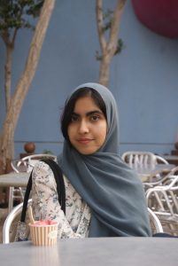 A young woman wearing a slate blue hijab and a white floral print blouse sitting at an outdoor cafe table. She is looking at the camera with a gentle smile. In front of her on the table is a small striped cup filled with pink gelato or ice cream, and the outdoor setting features seating, thin trees, and a blue wall in the background.