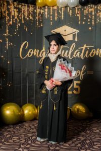 A graduate wearing a black academic gown, mortarboard, light beige headscarf, and a personalized stole holding a large bouquet of pink roses. She is standing in front of a celebratory dark backdrop that features golden script reading "Congratulations Class of '25", a graduation cap icon, and gold and black balloons along the top and floor.