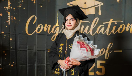 A graduate wearing a black academic gown, mortarboard, light beige headscarf, and a personalized stole holding a large bouquet of pink roses. She is standing in front of a celebratory dark backdrop that features golden script reading "Congratulations Class of '25", a graduation cap icon, and gold and black balloons along the top and floor.