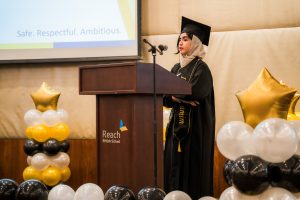 A graduate wearing a black academic gown, mortarboard, light beige headscarf, and a personalized stole standing behind a wooden podium speaking into a microphone. The podium features the "Reach British School" logo. The stage is decorated with gold star balloons and black, white, and yellow balloon arrangements, with a presentation screen in the background displaying the words "Safe. Respectful. Ambitious."