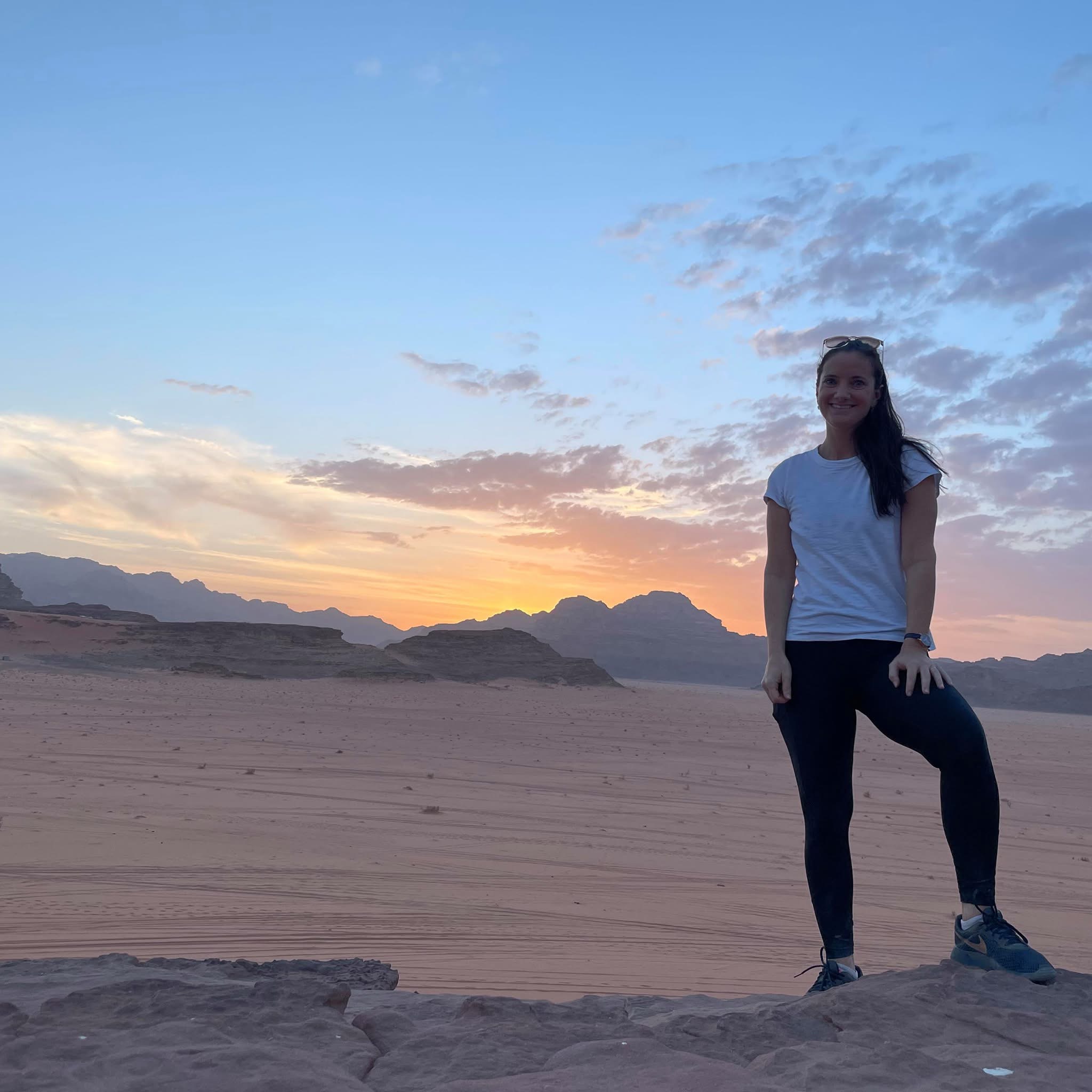 Image of Nicole standing on the right smiling on a rocky ledge in a vast desert landscape during sunset. She wears a short-sleeved white t-shirt, dark leggings, and sneakers, posing with one foot slightly elevated on a rock. In the background, low mountain ranges silhouette against a pale blue and orange twilight sky.