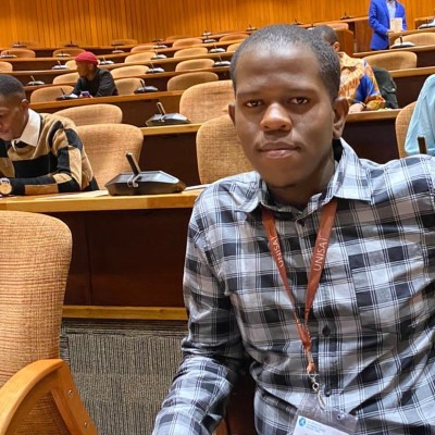 Portrait of a Nkeshimana wearing a grey plaid button-up shirt and a lanyard, seated in an auditorium or conference hall with rows of empty desks and chairs behind him.