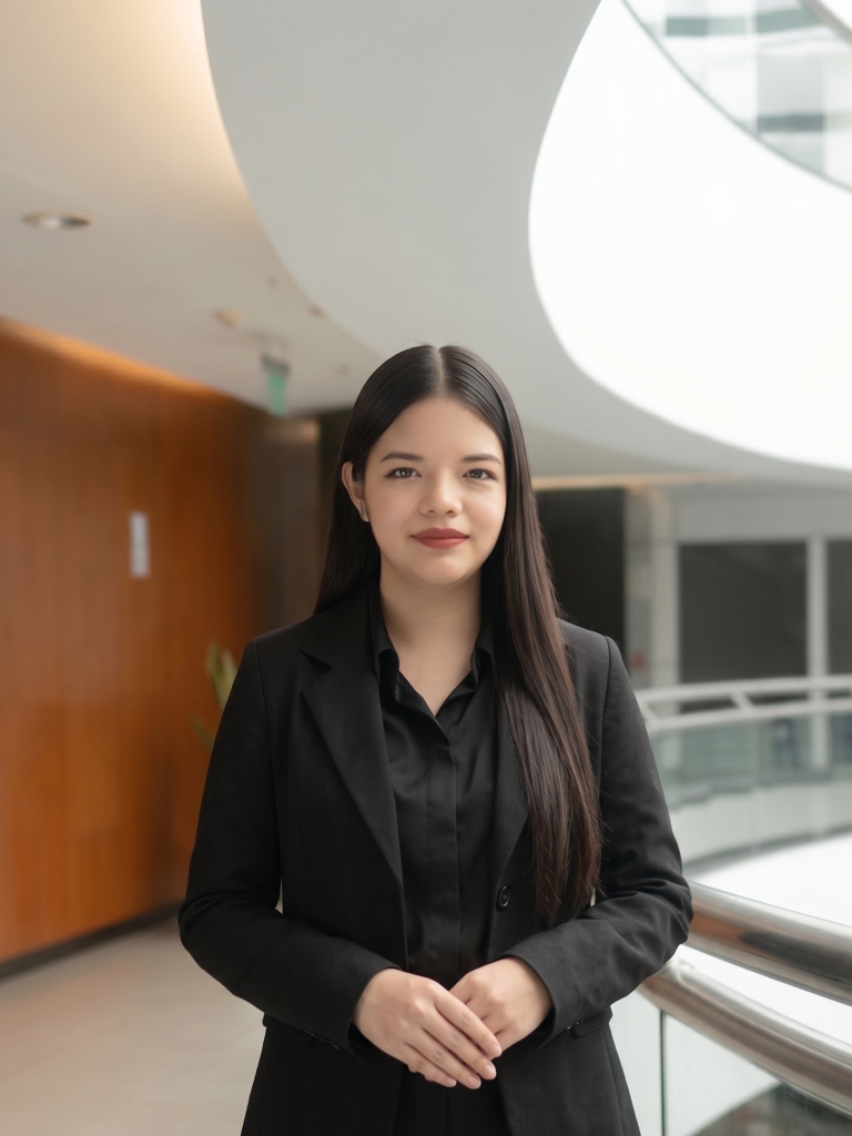 Professional portrait of Jasmin with long dark hair, wearing a black blazer and shirt, standing with her hands clasped in a modern indoor atrium.