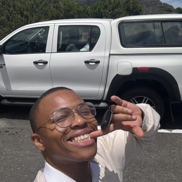 A smiling young man with round glasses holds up car keys for a selfie, standing outdoors in front of a white truck and scenic mountains.