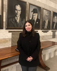 A young woman with fair skin and medium-length brown hair smiles at the camera with her hands clasped in front of her. She is wearing gray trousers, a black jacket, and hoop earrings. Behind her are a row of black and white portraits of men in formal attire, hanging in wooden frames on a stone wall.